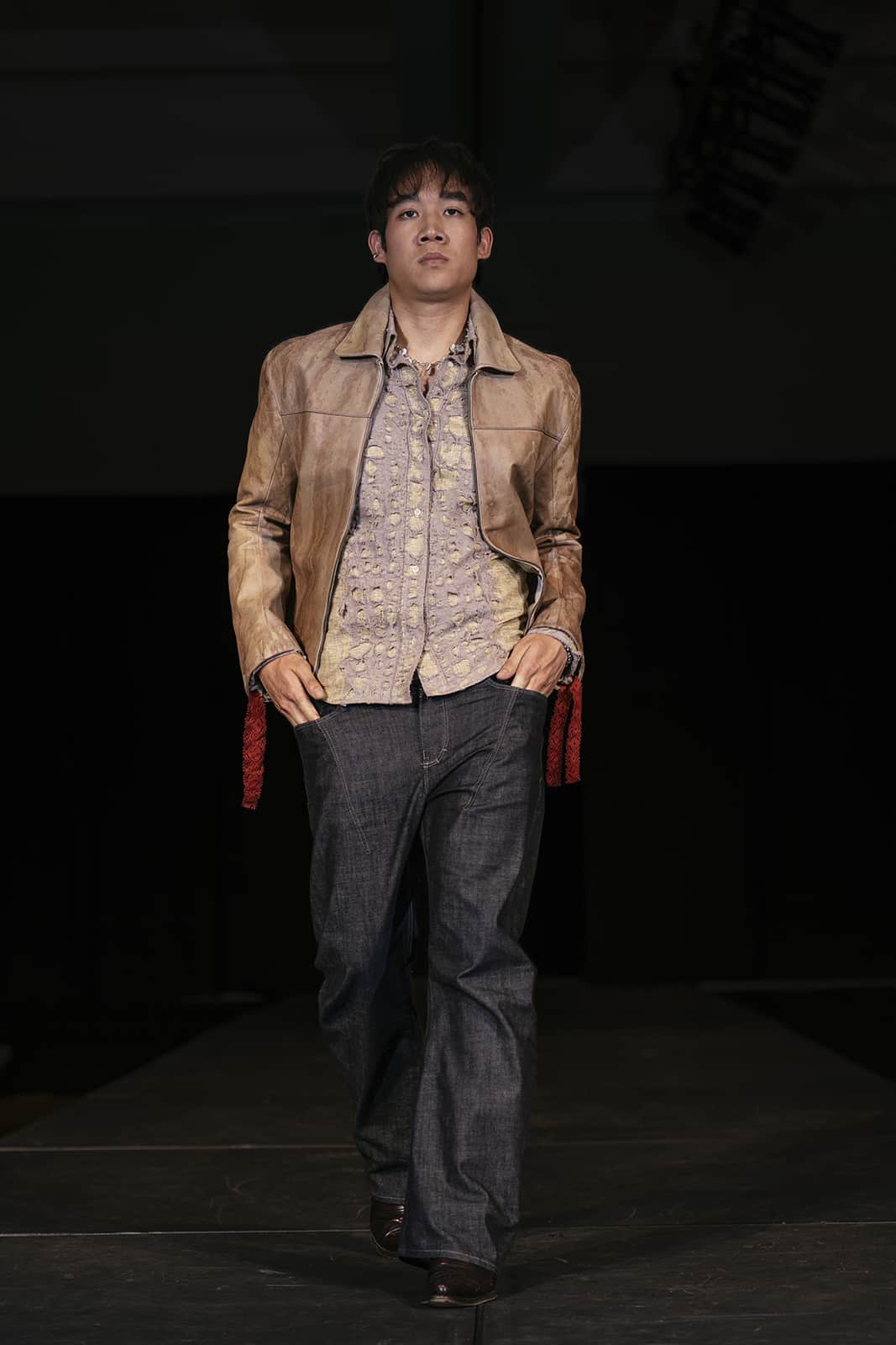 Model on the runway in jeans, a button down shirt and a brown jacket.