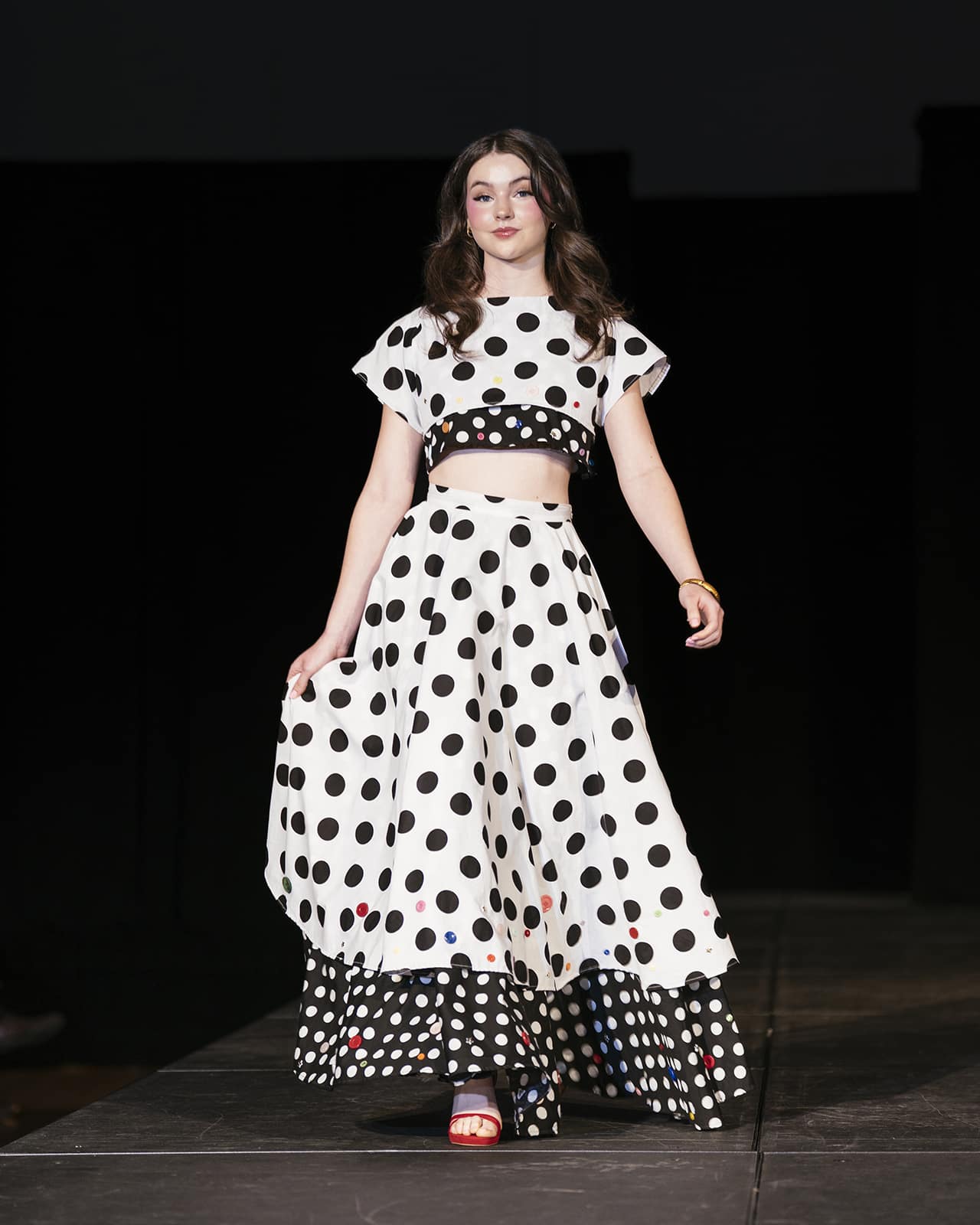 Model on the runway wearing a white with black polka dot two piece outfit with a top and a skirt.