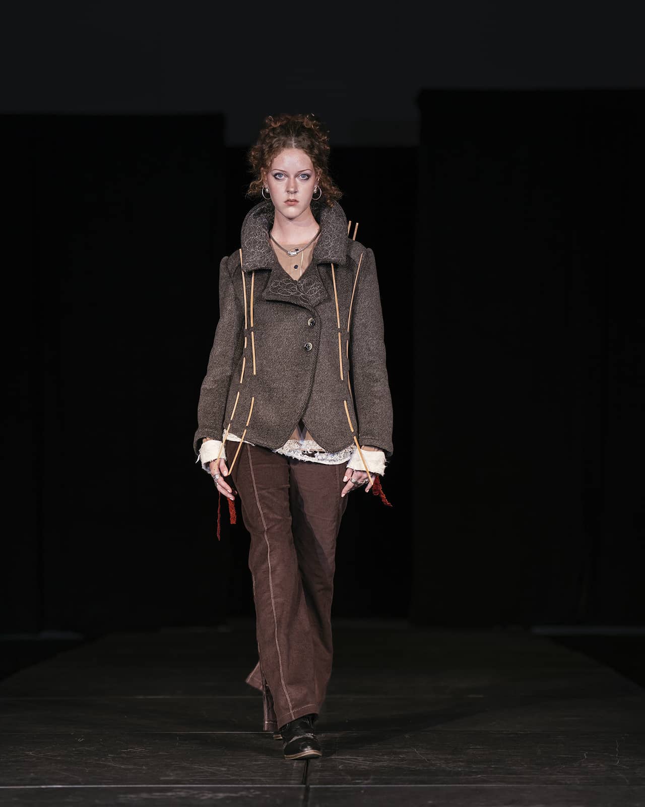 Model on the runway wearing brown pants, a white shirt and a brown jacket.