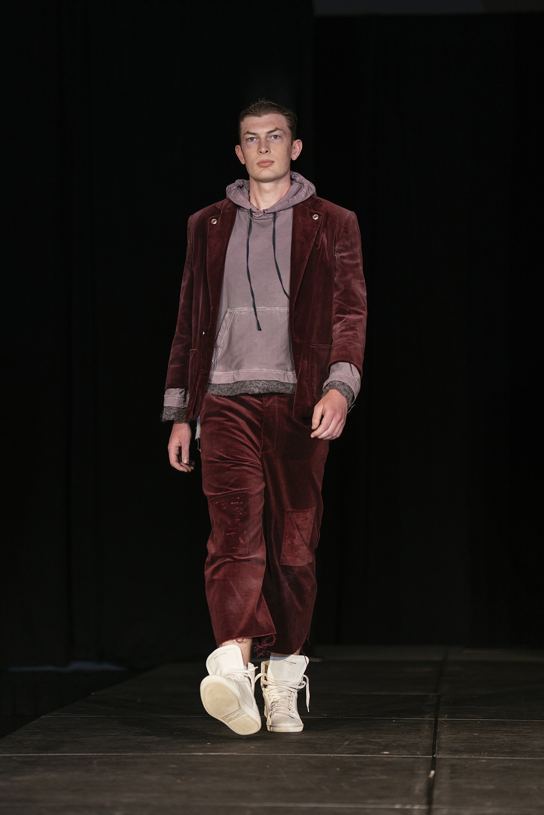 Model on the runway wearing crimson pants, a brown hoodie and crimson jacket.