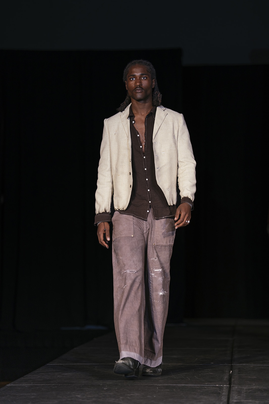 Model on the runway wearing brown pants, a brown button down shirt and white jacket.