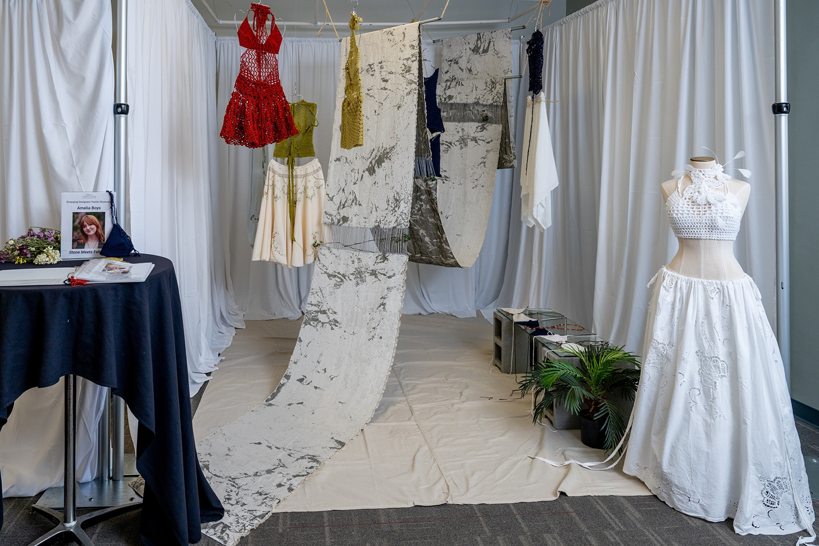 A photo of a room with one dress form with a white garment, other garments hanging from the ceiling as well as a long tapestry.