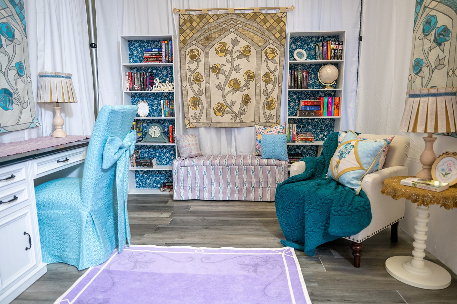 A photo of a room set-up with two bookcases flanking a wide chair, a tapestry with flowers above it, a white desk with blue chair and other textiles.