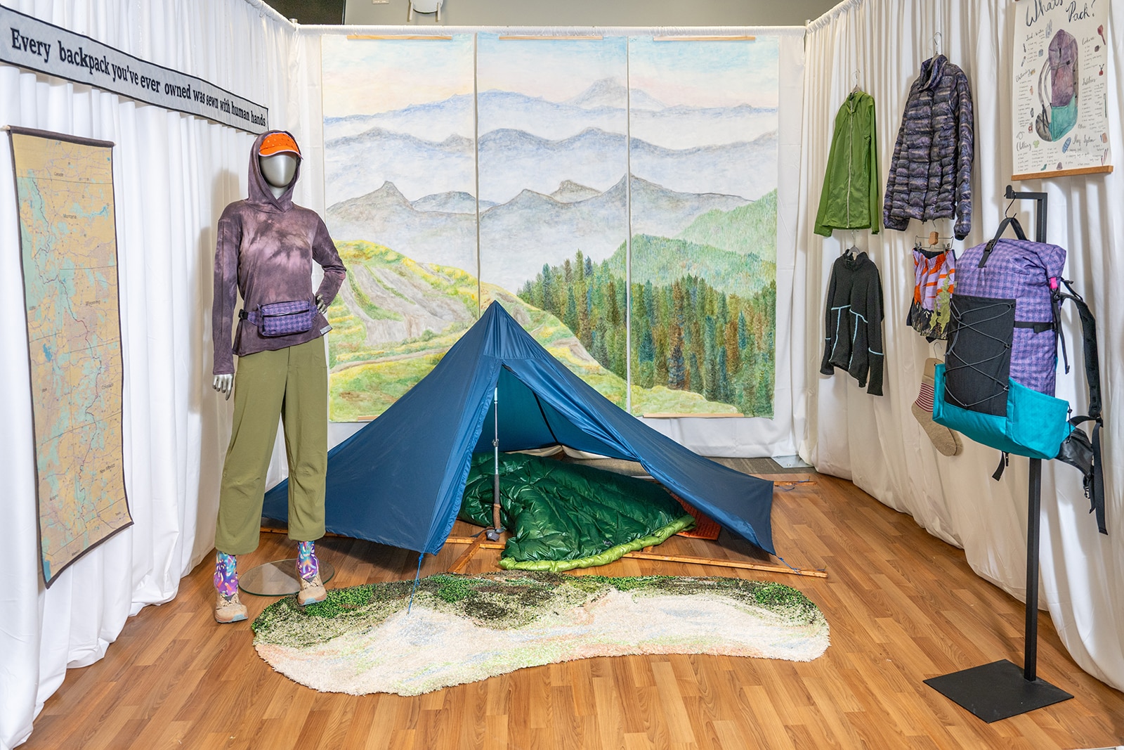 Photo of a room set-up to look like camping with a mannequin wearing green pants and a purple shirt, a teal tent, a mountain scene in the background and other garments in the room.