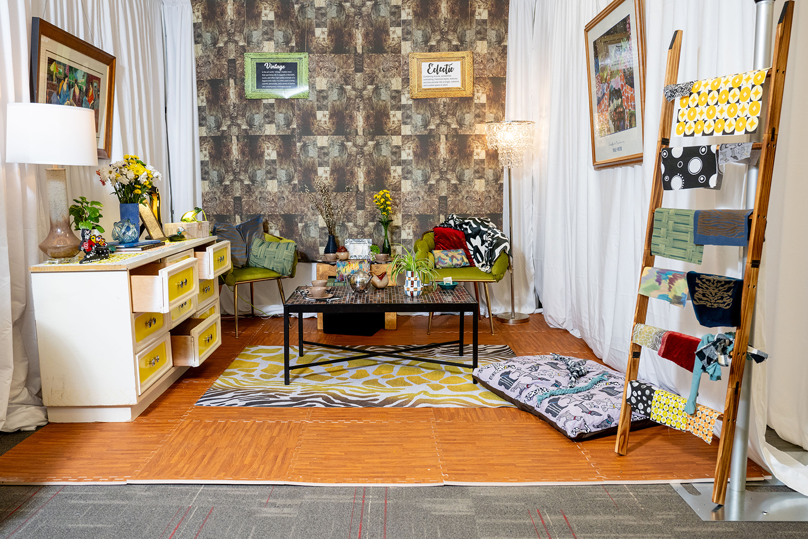Photo of a room with a wooden ladder with textiles on it, a desk off to the side with a lamp, a sitting area with a coffee table.