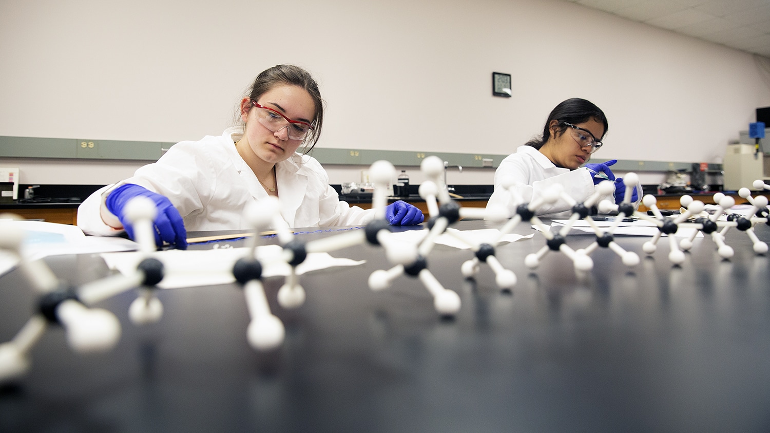 Two students wearing lab coats and safety glasses work in a lab.