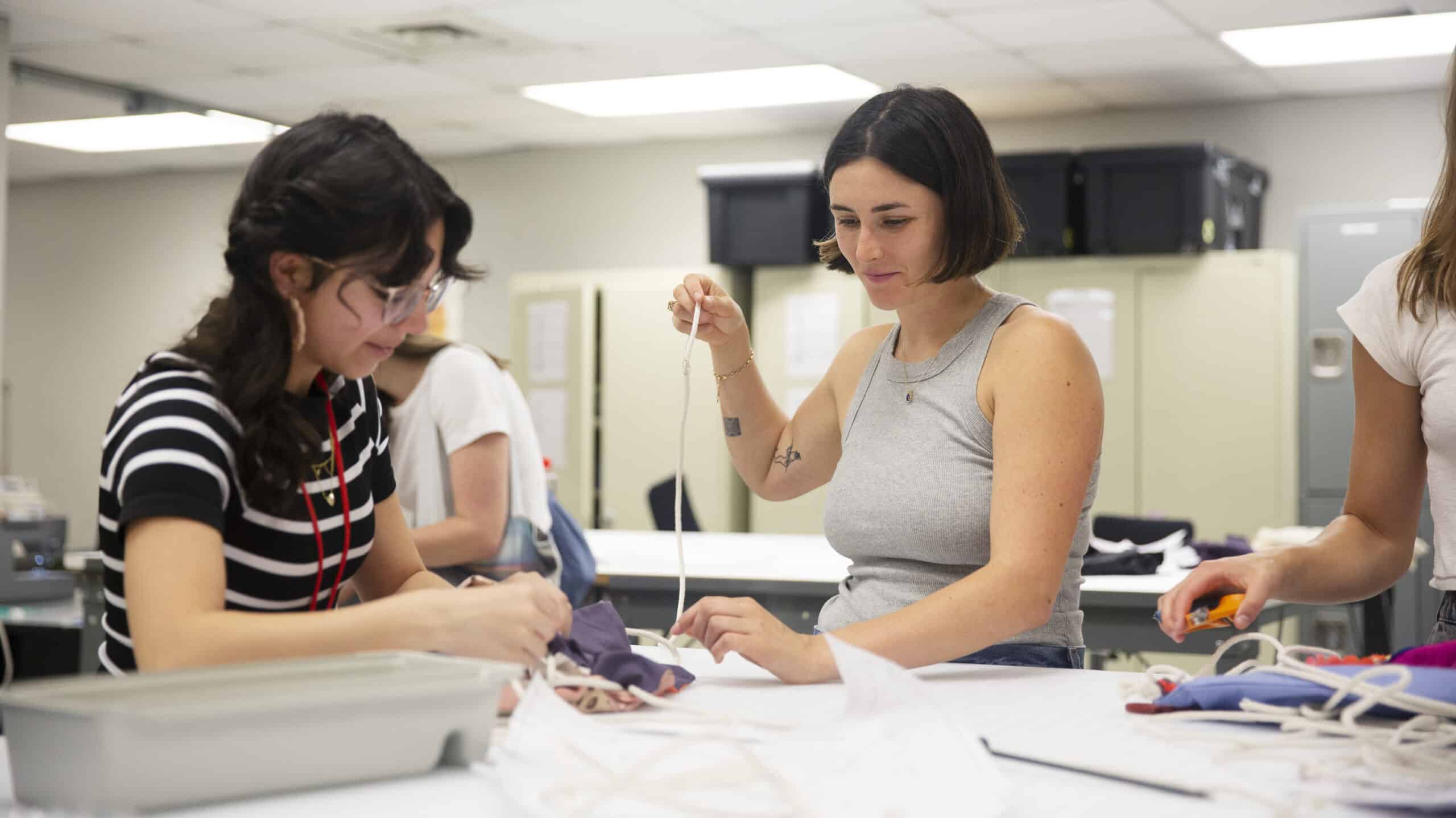 Two students work at a table. One student is pulling thread through a piece of fabric.