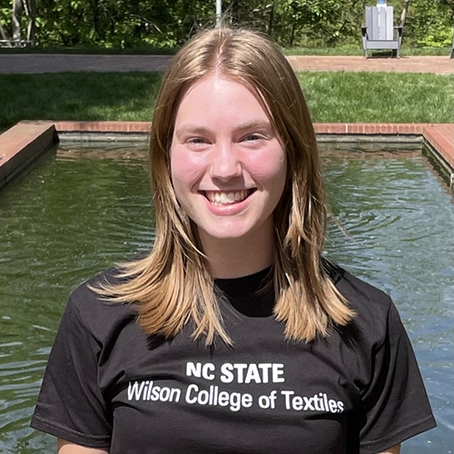Photo of a woman in a black t-shirt with a fountain behind her.