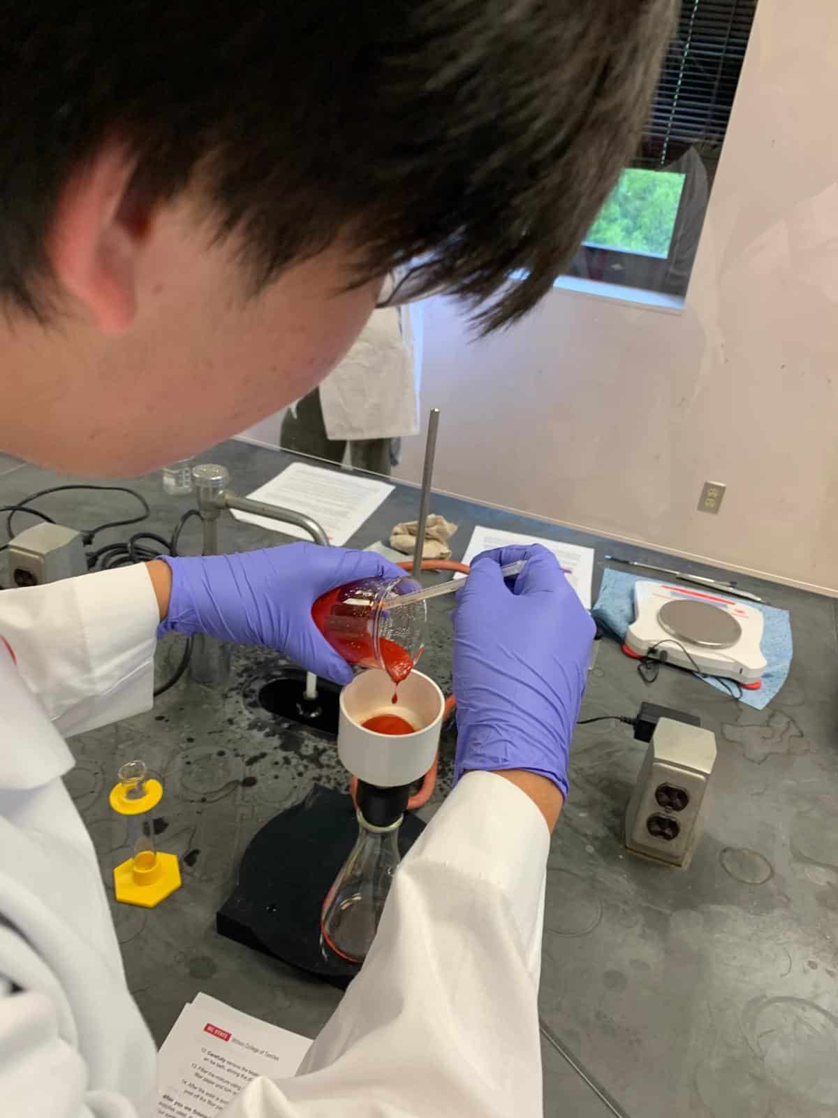 A student pouring a reddish semi-solid into a container in a lab environment
