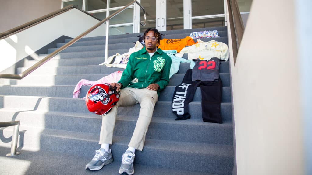 Teshaun Smith sits on a staircase with his designs and his football jersey with his right hand. nd him. He holds a football helmet with h