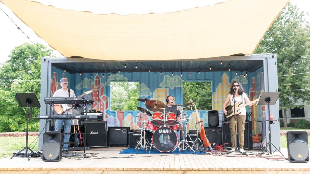 A band plays on an outdoor stage made out of a deck and an old shipping container. The shipping container has a portrait of a city painted on it, as well as string lights. The band is composed of a guitarist and keyboardist (left) and drummer (center) and guitarist and vocalist (Kate Annett-Hitchcock, right). 