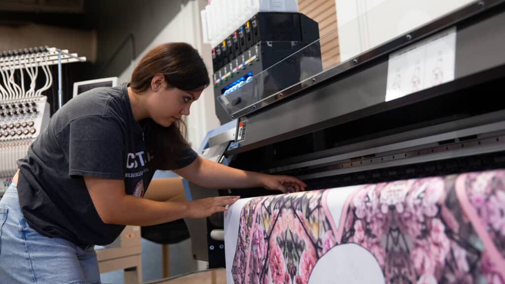 To the left of the frame, Annie Hoyt leans over a pink fabric sample to adjust its position. To the right, the fabric is draped over a printing machine.