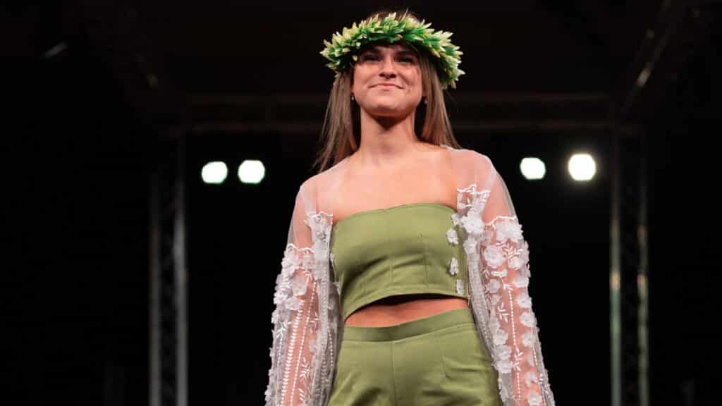 Model stands on a runway and is visible from the waste up. They wear a "crown" of greenery on their head and a green linen crop top and shorts matching "set." On top of that, they wear a sheer shawl embellished with white flowers. 