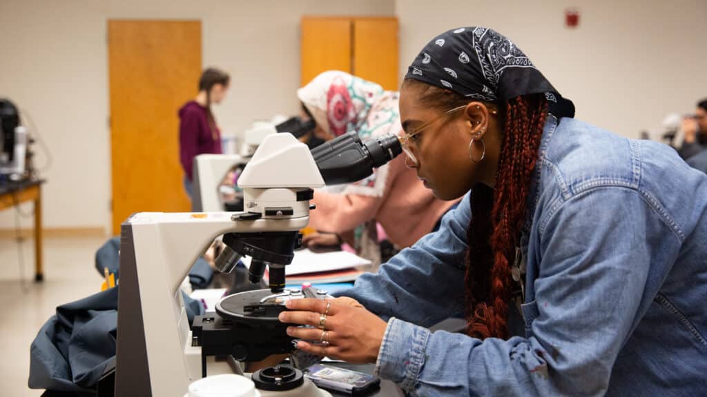 Two students look at a microscope. The one closest to the camera adjusts the plate. The one furthest from the camera looks into the lens.