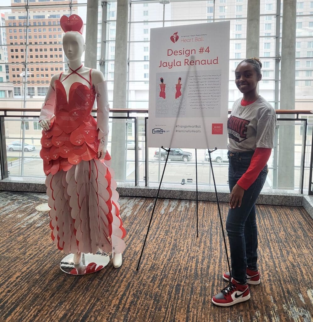 A student stands next to a mannequin dressed in a red gown and matching red crown made entirely from paper hearts. A sign reads: “Design #4 Jayla Renaud”.