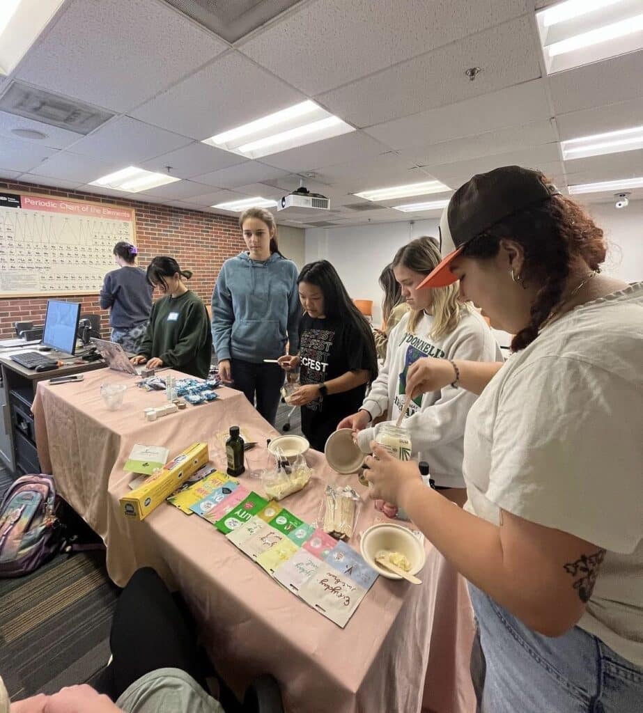 Six students gather around a table with materials for making homemade shea body butter. The table has a pink tablecloth and on top of it there are face masks, bowls, and other materials for the DIY. 