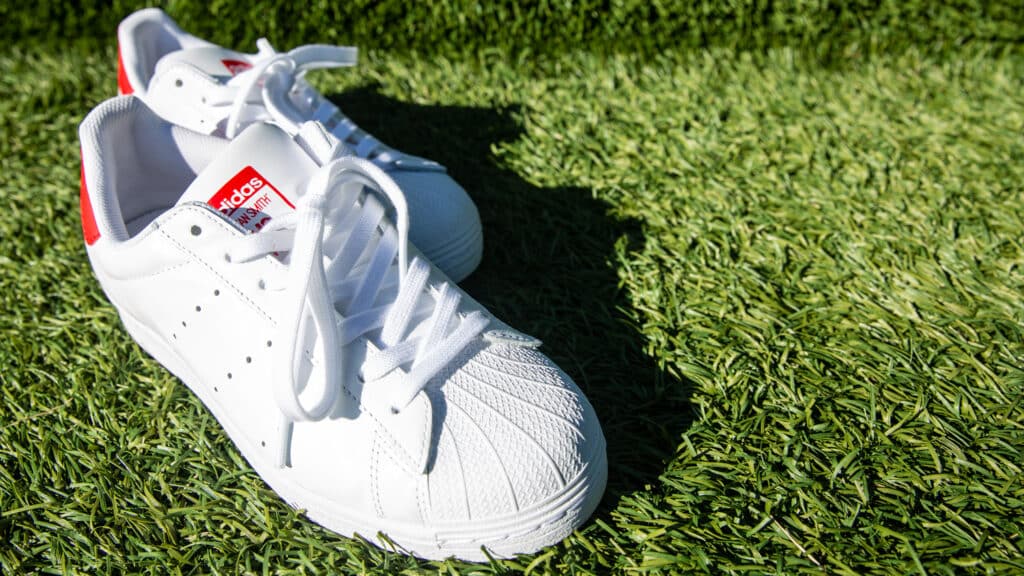 White Stan Smith Shell Toe sneakers in white with a bright red heel are laid out in a staggered fashion on synthetic turf. 