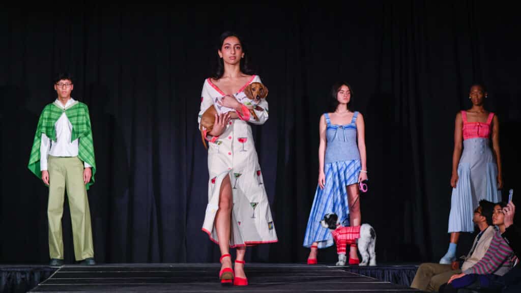 Four models on a raised black runway with a black curtain backdrop. The model second from left is walking down the runway, while the rest stand on the back end of the stage. The model on the farthest left wears a white dress shirt, a green plaid cape and green dress pants. The model walking down the runway wears a khaki, longsleeved dress shirt with pink buttons, pink piping and martini glasses embroidered all over the dress; she carries a dachshund wearing a matching jacket. The model second from the right wears a light blue sleeveless sweater with blue striped straps, as well as a blue, plaid asymmetrical skirt; that model holds a dog on a leash with a pink plaid sweater. The model on the farthest right wears a pink sleeveless crop top and a blue pleated skirt with fabric-covered buttons. 