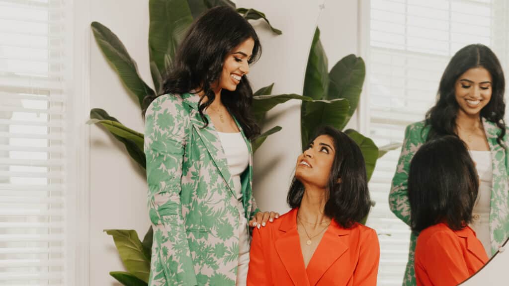 A photo of Ritika and Niki Shamdasani. Ritika stands with her hand on the shoulder of Niki, who is sitting to the right of her. Both are looking at each other and smiling. They are in a bright, white walled room with a green plant behind them. A mirror is behind Niki. Ritika wears a green and pink floral blazer. Niki wears a tangerine orange blazer.