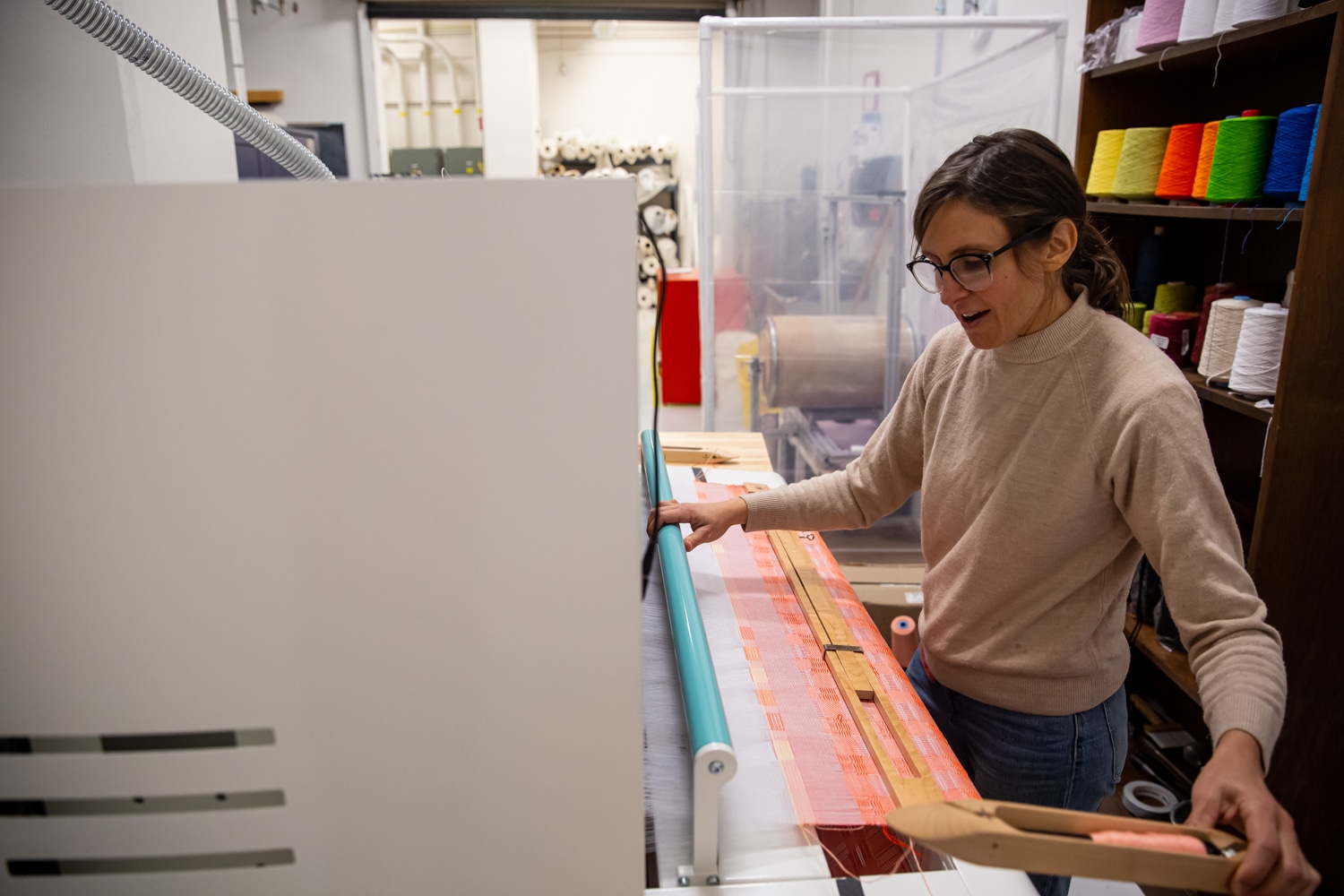 Kate Nartker uses TC2 loom in the Wilson College of Textiles lab. Behind her, a bookshelf with spools of colorful thread can be seen. The beginnings of the weave are visible: a bright orange geometric pattern.