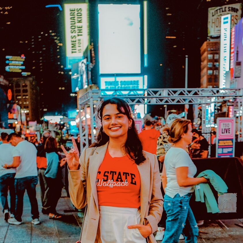 Ankitha Gurram stands in Times Square at night. She poses with her "wolves up" and wears a red NC State t-shirt with a tan blazer.