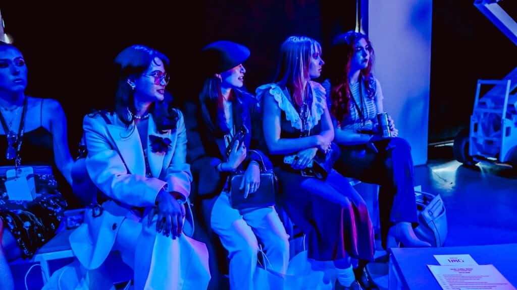 Photo of Ankitha Gurram and other students sitting on a bench inside the headquarters for New York Fashion Week. All are in blue light and looking right towards a stage (not shown) where a runway show is about to begin.