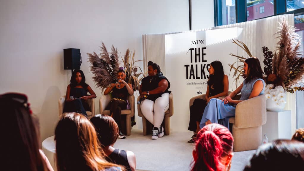 On a raised stage, five panelists sit in mid-century style chairs and speak to an audience. 