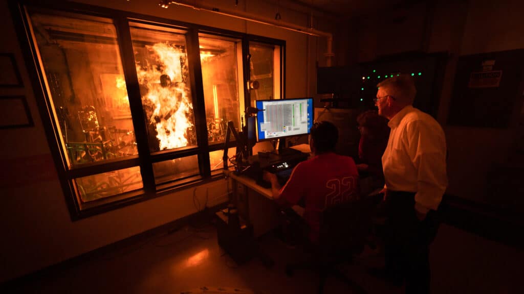 Student and faculty watch Pyroman manikin on fire, testing garment flammability and thermal protection.