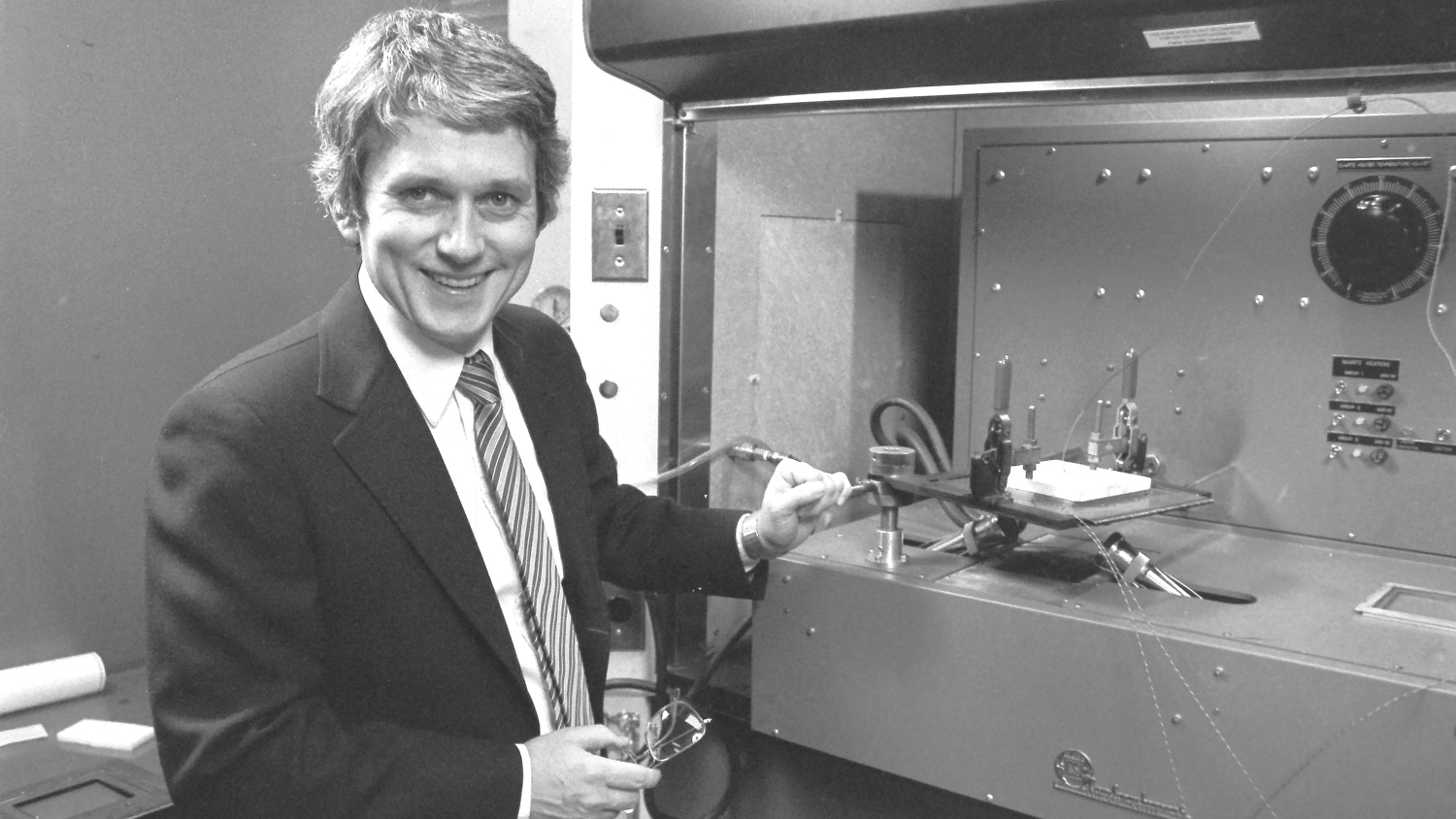 Black and white photo of Roger Barker. He wears a suit and tie and stands smiling and looking at the camera in a lab.