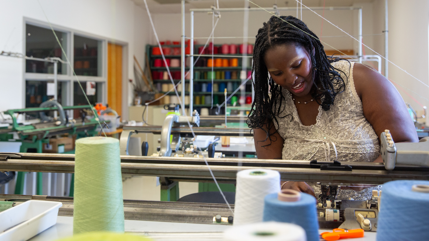 Maddy Moore works in a textile workshop, adjusting a knitting machine. Various spools of thread in different colors are on the table in front of her. Shelves with more colorful spools line the back wall. The room is brightly lit.