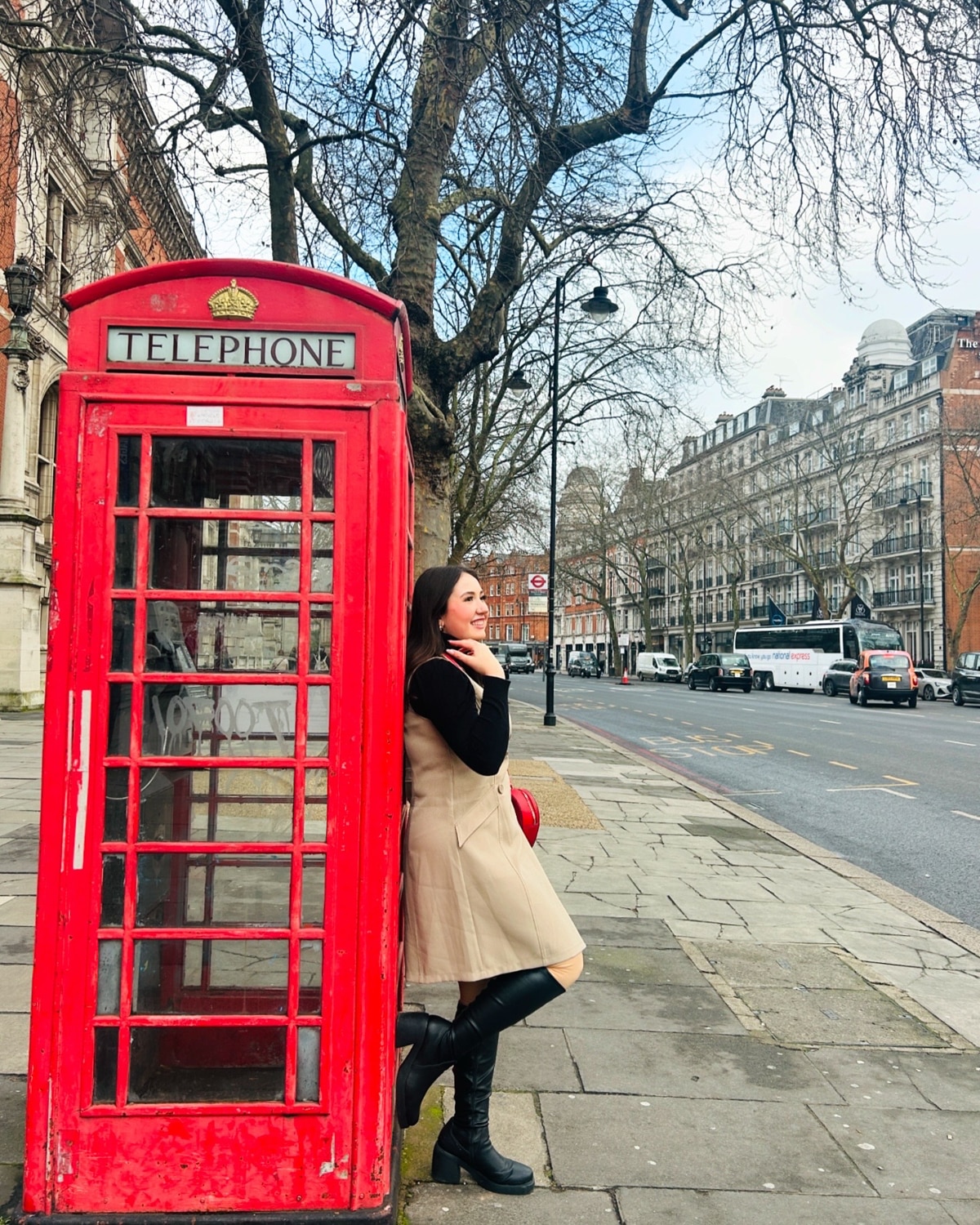 Lainey Volz, wearing a khaki dress and black boots leans against a classic red British telephone booth on a city street lined with trees and historic buildings, with cars and buses in the background.