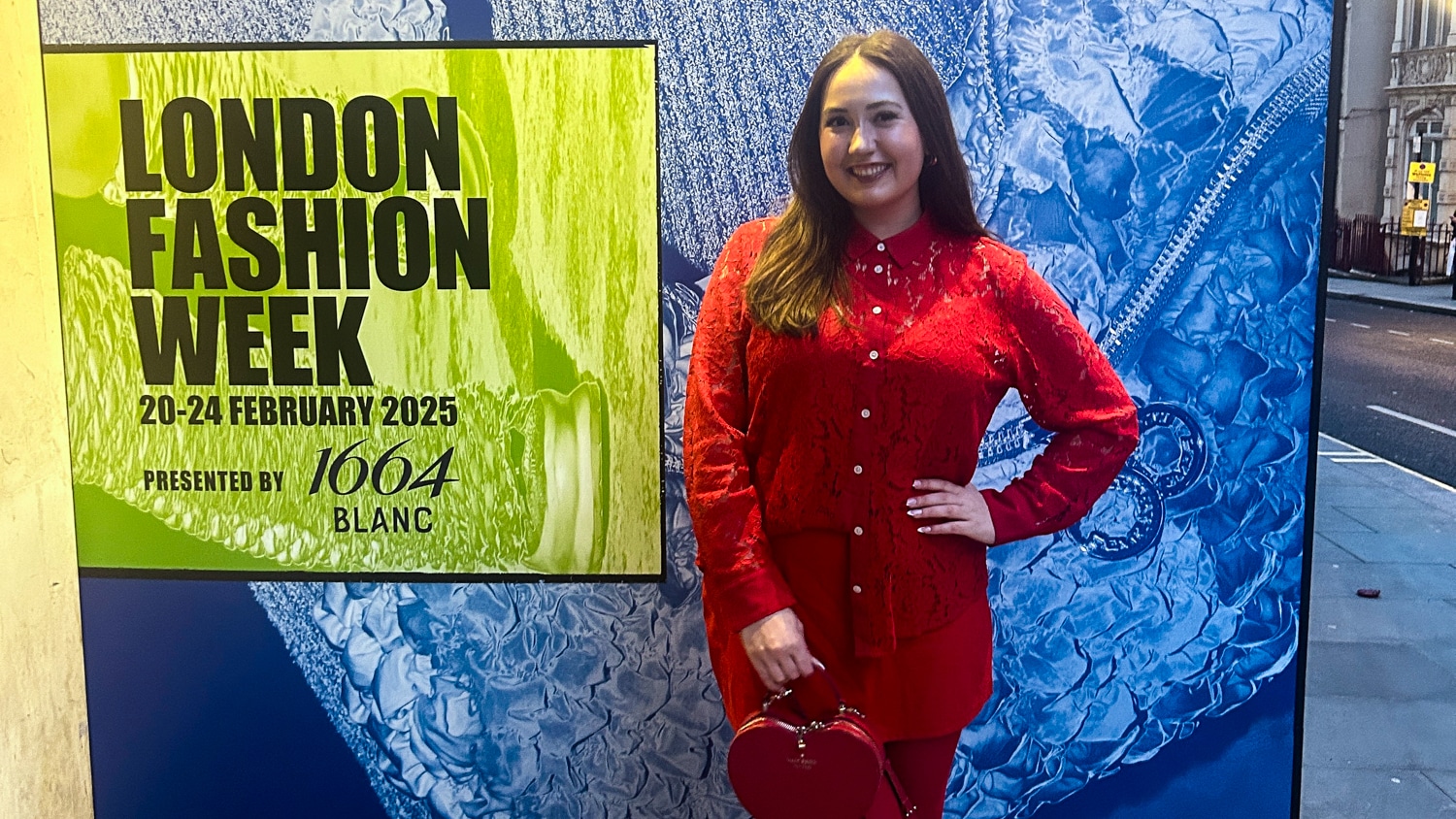 Lainey Volz wears an all red outfit and stands smiling next to a London Fashion Week 2025 sign on a city street.