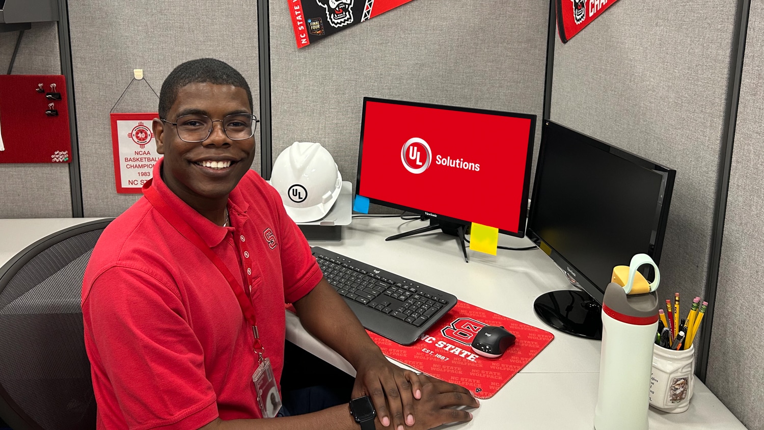 Darian Boyd sits at a desk in an office cubicle with a UL Solutions logo on the computer screen, surrounded by NC State decorations and office supplies. He is smiling and wears glasses and a red polo.