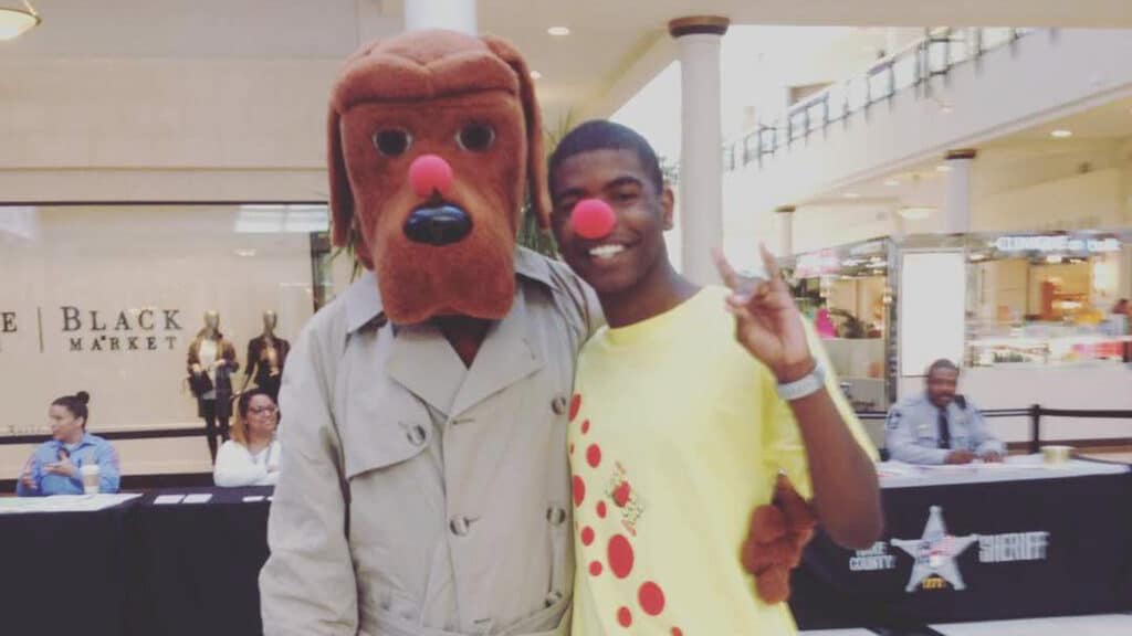  Darian Boyd in a yellow shirt with a red clown nose stands next to someone in a large brown dog costume wearing a trench coat, both smiling and posing in a mall with people seated at tables in the background.


