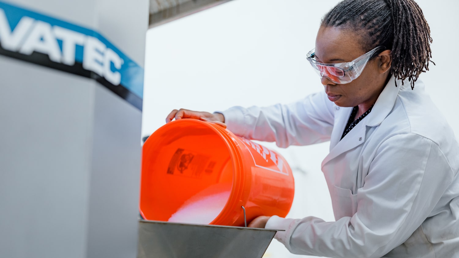 Ericka Ford, wearing safety goggles and a lab coat, pours white granules from a large orange bucket into equipment in a laboratory.
