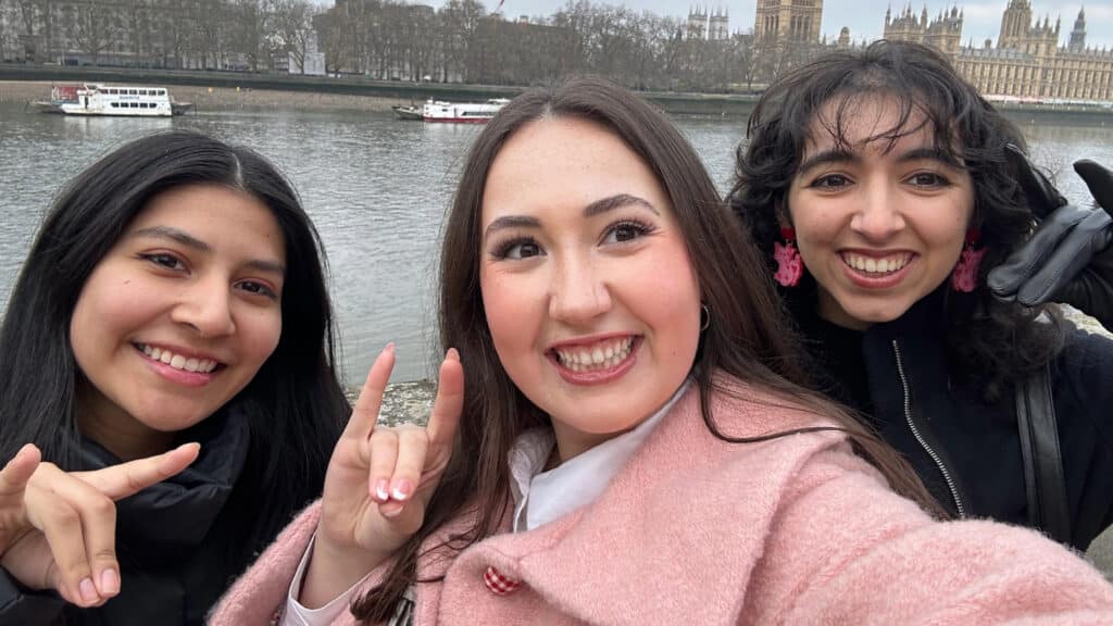 Three students smiling at the camera with the London skyline in the background.
