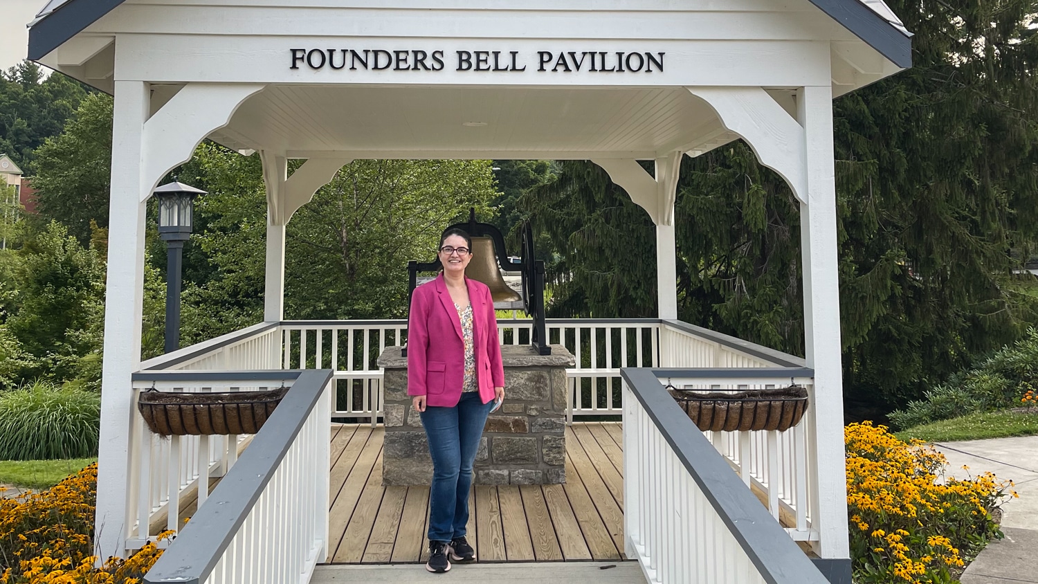 Nimet Degirmencioglu stands smiling under the Founders Bell Pavilion, surrounded by greenery and yellow flowers.