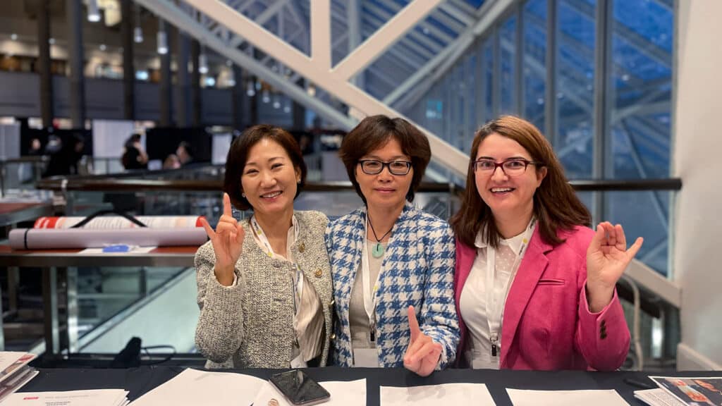 Nimet Degirmencioglu, Yingjiao Xu and Ellie Jin (from right to left) sit at a table indoors, smiling at the camera and making wolves up signs. They are dressed in business attire and there are papers and a phone on the table. A modern glass and steel structure is in the background.