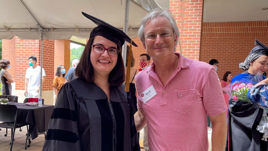 Nimet Degirmencioglu (in a graduation cap and gown) smiles next to David Hinks at an outdoor ceremony. Other people and a brick building are visible in the background.