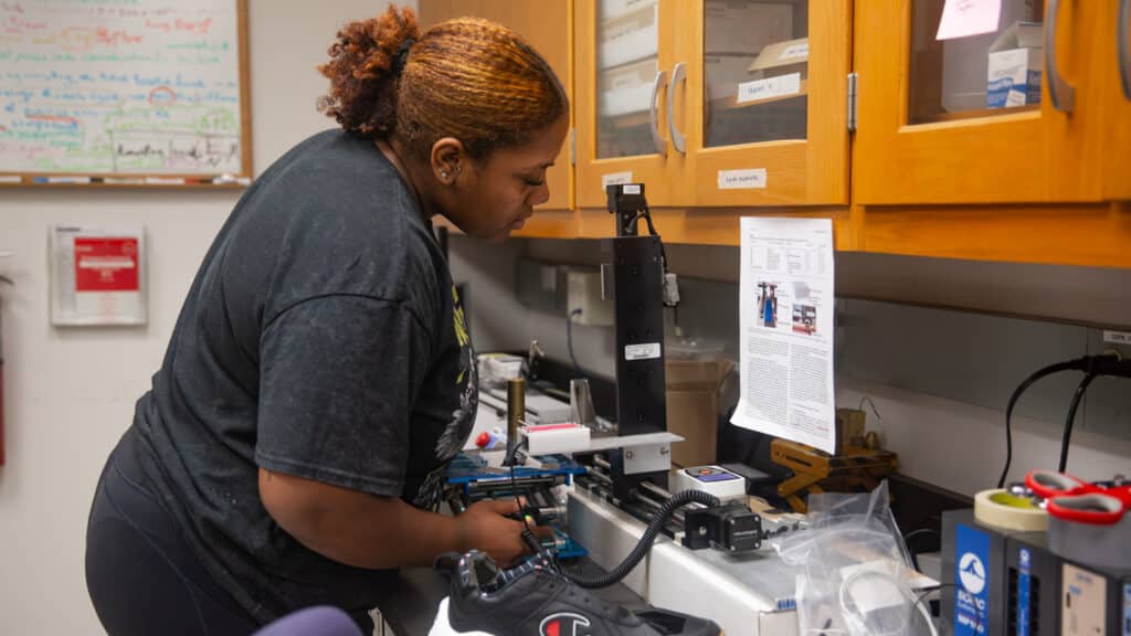 Andrianna Hodges works in a laboratory, using equipment on a counter with various scientific tools, cabinets, and papers visible around them.