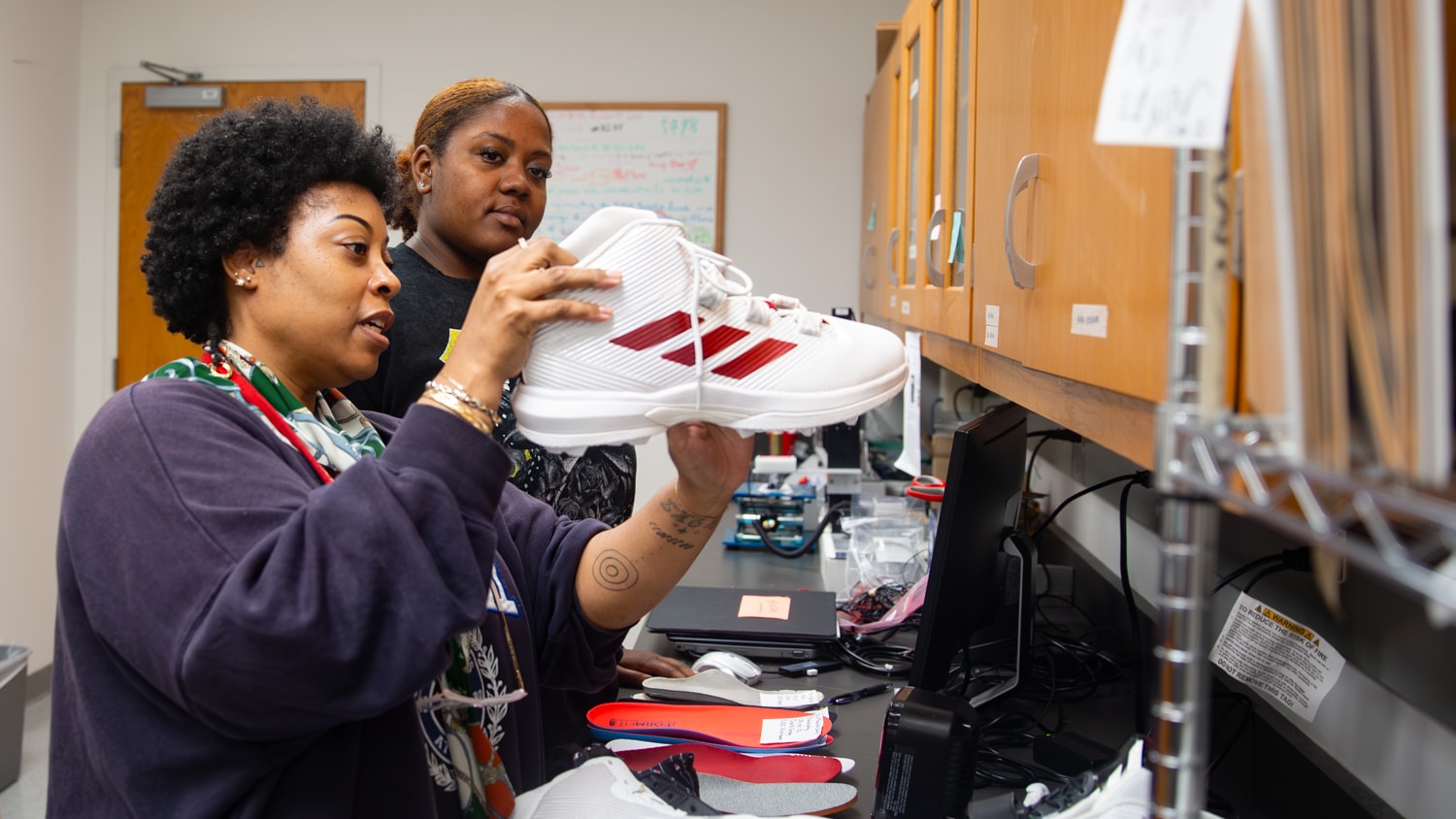 Kedena Henriques-Thompson and Andrianna Hodges examine a white athletic shoe with red stripes in a lab space. One woman holds the shoe up while the other observes.