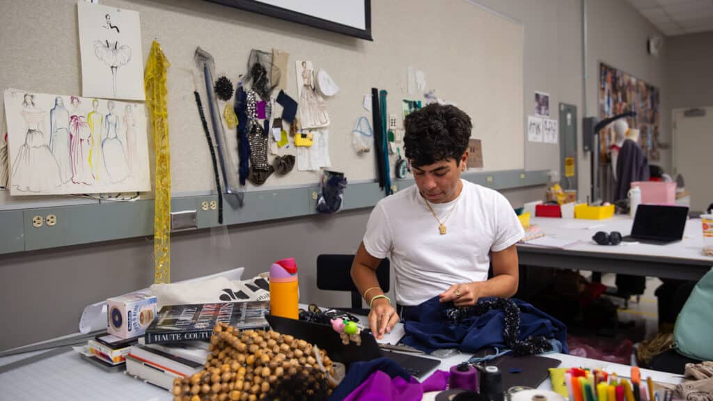 A person in a white shirt works with fabric at a table in a fashion studio, surrounded by sewing materials, books, sketches, and supplies pinned to a wall behind them.