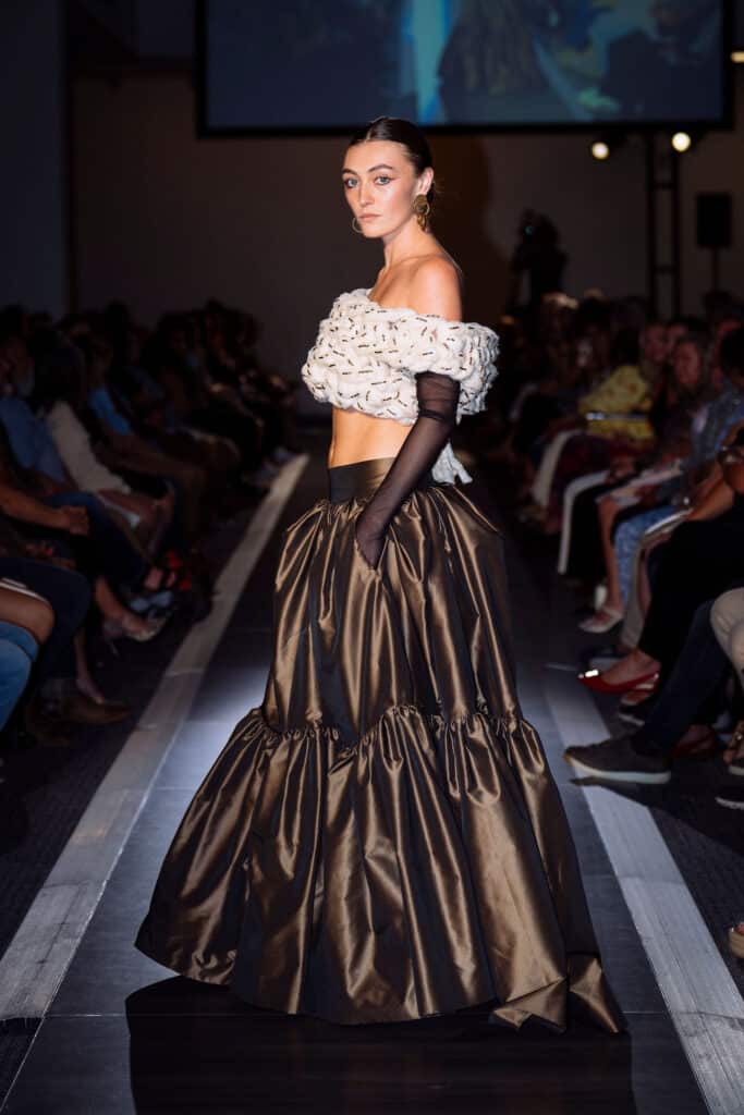 A model walks down a runway wearing a white textured off-shoulder top, long black gloves, and a voluminous brown satin skirt. The audience is seated on both sides, and the lighting highlights the model.