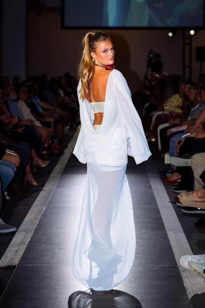 A model in a sheer white gown with an open back walks on a runway, looking over her shoulder at the camera. The audience sits on both sides watching the fashion show.
