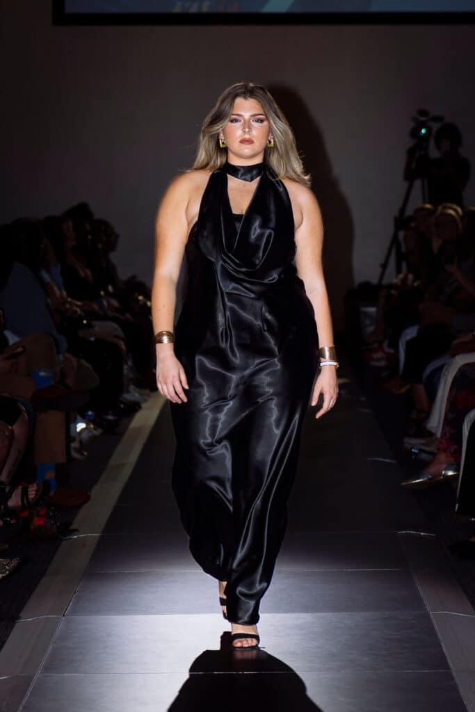 A model with long blonde hair walks down a runway wearing a sleeveless, black satin dress and black heels, accessorized with gold bracelets and earrings. The audience sits on both sides of the runway.