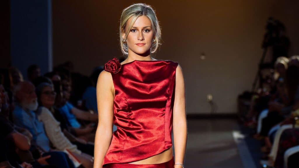 A model walks down a runway wearing a shiny red sleeveless dress with a rose detail on the shoulder. The audience is seated on both sides, and the background is softly lit.