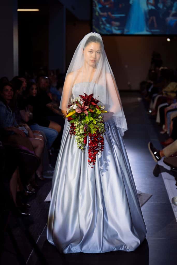 A bride in a silver strapless gown and veil holds a cascading bouquet of red, yellow, and green flowers while walking down a runway, with an audience seated on both sides.