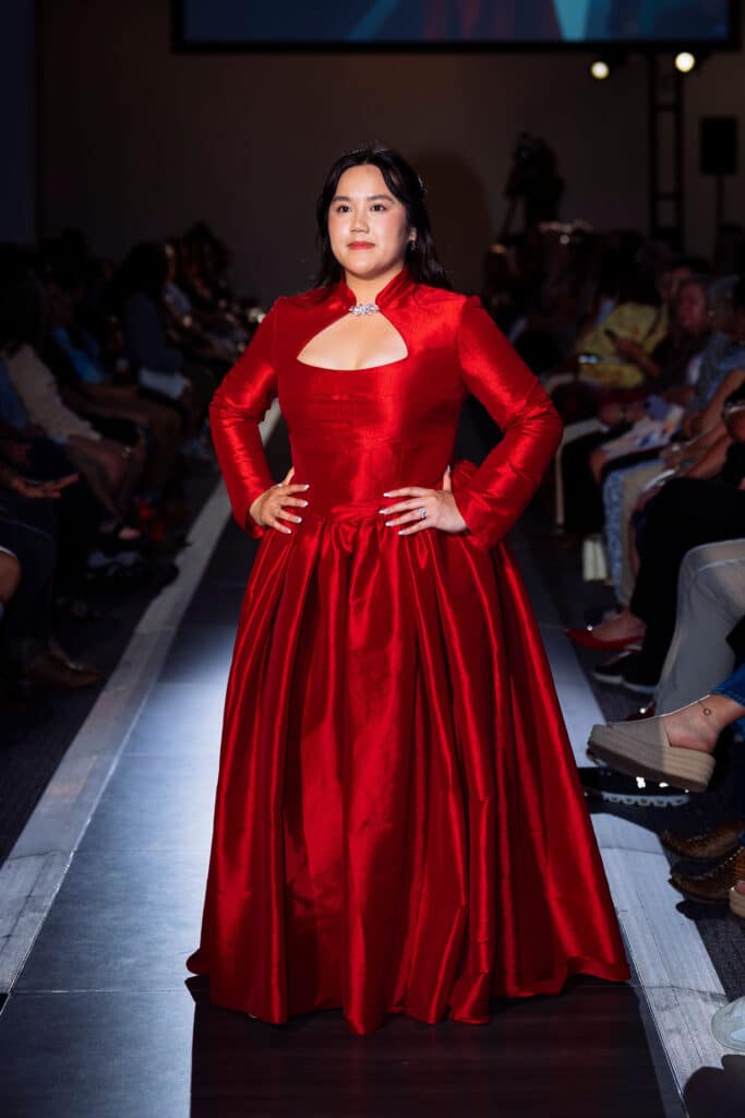 A woman confidently walks down a runway wearing a long, elegant, bright red gown with long sleeves as an audience watches from both sides.