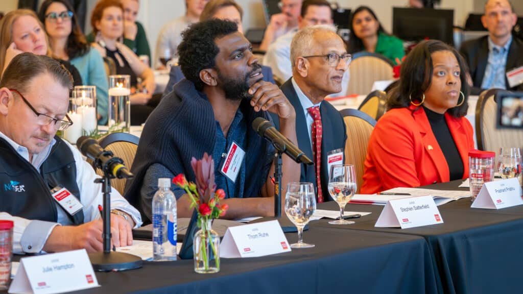Stephen Satterfield and three other people sit at a table with microphones, nameplates and drinks, attentively listening during an event. Audience members are visible in the background.