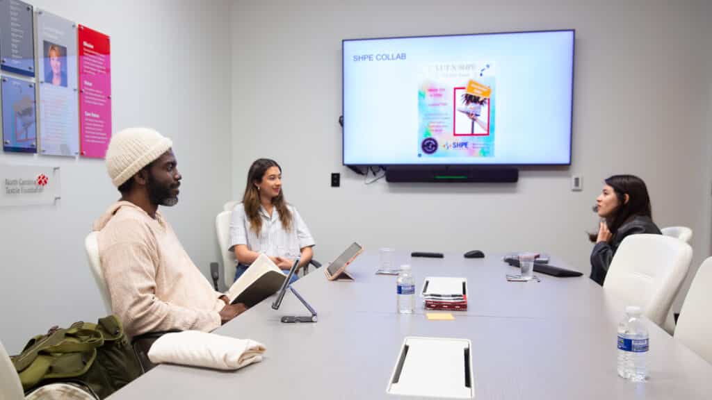 Stephen Satterfield and two other people sit around a conference table in a meeting room, with notebooks and tablets. A presentation is displayed on a screen at the front of the room.